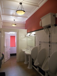 A restroom with white tiled walls and a red accent wall features two sinks with mirrors and three urinals. The ceiling is paneled with two hanging light fixtures, and the floor appears to be tiled. There is a window covered by a red curtain and an older-style toilet cistern mounted high on the wall.