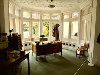 A vintage office setup with a large wooden desk facing a bay window. The desk is adorned with a few items, including a lamp and a hat placed on a coat stand. The window features decorative stained glass panels above clear glass panes, allowing natural light to illuminate the room. The walls and ceilings have intricate molding details, and a single chair is positioned near the desk. A green carpet covers the floor, creating a cozy yet formal atmosphere.