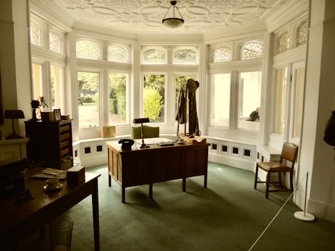 A vintage office setup with a large wooden desk facing a bay window. The desk is adorned with a few items, including a lamp and a hat placed on a coat stand. The window features decorative stained glass panels above clear glass panes, allowing natural light to illuminate the room. The walls and ceilings have intricate molding details, and a single chair is positioned near the desk. A green carpet covers the floor, creating a cozy yet formal atmosphere.