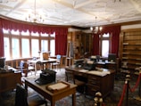 A vintage office with wooden furniture, typewriters, and an ornate ceiling. Red draped curtains frame large windows that let in natural light. The room features multiple desks and chairs, with shelves lining the walls and various office supplies placed around the room.