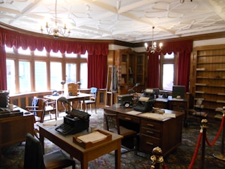 A vintage office with wooden furniture, typewriters, and an ornate ceiling. Red draped curtains frame large windows that let in natural light. The room features multiple desks and chairs, with shelves lining the walls and various office supplies placed around the room.