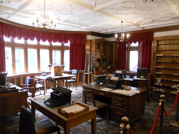 A vintage office with wooden furniture, typewriters, and an ornate ceiling. Red draped curtains frame large windows that let in natural light. The room features multiple desks and chairs, with shelves lining the walls and various office supplies placed around the room.
