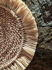 A woven straw mat or placemat is partially visible against a textured, irregular stone surface. The straw fibers are thickly layered and arranged in a circular pattern with frayed edges, showcasing a rustic and natural aesthetic.