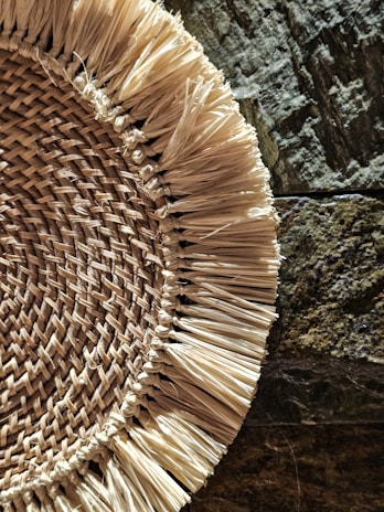 A woven straw mat or placemat is partially visible against a textured, irregular stone surface. The straw fibers are thickly layered and arranged in a circular pattern with frayed edges, showcasing a rustic and natural aesthetic.