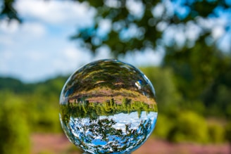 A crystal ball reflects an inverted image of a lush green landscape with a bright blue sky and scattered clouds. The spherical object creates a unique perspective, highlighting the greenery and sky with vibrant clarity. The background is softly blurred, showcasing a deep depth of field focused on the crystal ball.
