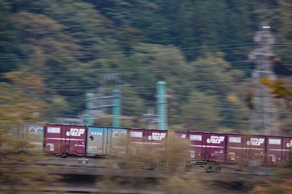 A moving freight train is captured in motion, with bright cargo containers prominently displayed against a backdrop of blurred greenery and utility poles. The train is composed of purple and blue containers, each bearing distinctive logos and text.