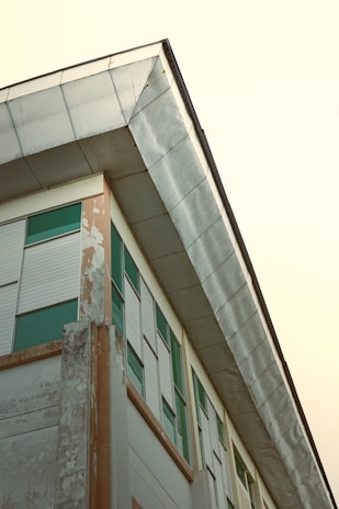 A building corner with large metal siding extends upwards, featuring rows of green-tinted windows and patches of weathered, peeling paint on the surface.