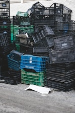 A large pile of stacked plastic crates, mostly black and a few green and blue, are arranged haphazardly against a light-colored wall. Some crates are overturned, creating a chaotic appearance. A piece of cardboard lies on the ground in front of the stack.