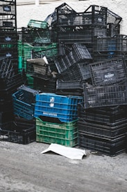 A large pile of stacked plastic crates, mostly black and a few green and blue, are arranged haphazardly against a light-colored wall. Some crates are overturned, creating a chaotic appearance. A piece of cardboard lies on the ground in front of the stack.