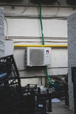 An outdoor air conditioning unit is mounted on a textured wall with various pipes and electrical conduits running above it. The unit is labeled GREE and is connected to a green hose. Below, several empty black plastic crates are stacked haphazardly on a rough concrete surface.