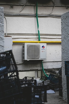 An outdoor air conditioning unit is mounted on a textured wall with various pipes and electrical conduits running above it. The unit is labeled GREE and is connected to a green hose. Below, several empty black plastic crates are stacked haphazardly on a rough concrete surface.