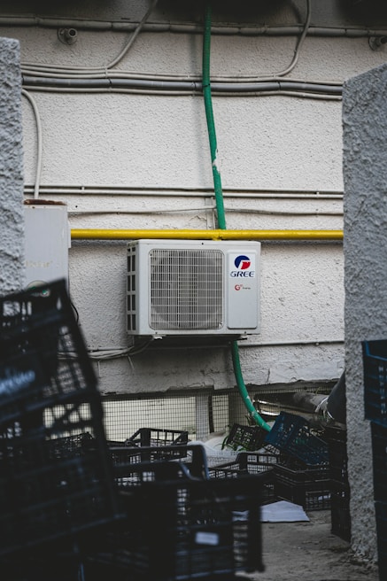 An outdoor air conditioning unit is mounted on a textured wall with various pipes and electrical conduits running above it. The unit is labeled GREE and is connected to a green hose. Below, several empty black plastic crates are stacked haphazardly on a rough concrete surface.