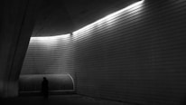 A dimly lit architectural space with angular and curved concrete walls. A single silhouette of a person stands near a vent or bench, creating a sense of solitude and introspection. The space is illuminated by a line of lights casting dramatic shadows.