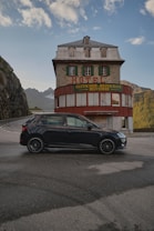 A vintage hotel building with a red and green color scheme stands against a backdrop of mountains. The hotel sign reads 'Gletscher-Restaurant Belvedere' and it is positioned on a winding mountainous road. A modern black car is parked in front of the hotel. The sky is partly cloudy with scattered clouds.