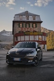 A black car is parked in front of an old stone building with the word 'HOTEL' prominently displayed above green window shutters. The building has a rustic appearance with a metal roof and is surrounded by mountainous terrain. The sky is partly cloudy with soft lighting, suggesting it might be early morning or late afternoon.