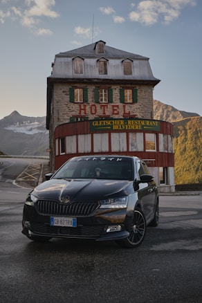 A black car is parked in front of an old stone building with the word 'HOTEL' prominently displayed above green window shutters. The building has a rustic appearance with a metal roof and is surrounded by mountainous terrain. The sky is partly cloudy with soft lighting, suggesting it might be early morning or late afternoon.