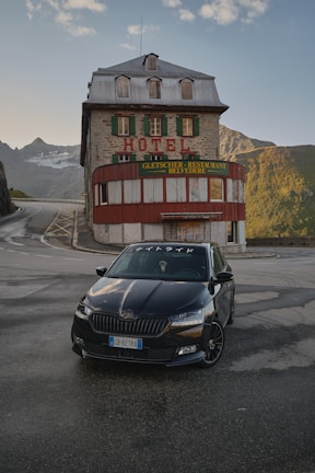 A dark-colored car is parked in front of an old stone hotel with green shutters and a red and green sign reading 'Gletscher Restaurant Belvedere.' The building is set in a mountainous area with winding roads, and the sky is partly cloudy with the light of either early morning or late evening.