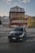 A dark-colored car is parked in front of an old stone hotel with green shutters and a red and green sign reading 'Gletscher Restaurant Belvedere.' The building is set in a mountainous area with winding roads, and the sky is partly cloudy with the light of either early morning or late evening.