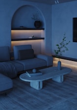 A modern living room with a minimalist aesthetic. The space features a sectional sofa with a throw blanket, accented by a minimalist coffee table with books and a decorative orb. Shelves are built into an alcove, displaying a potted plant and other small items, softly illuminated by warm lighting. The room includes neutral tones and natural textures, contributing to a calming atmosphere.
