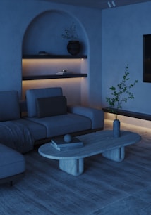 A modern living room with a minimalist aesthetic. The space features a sectional sofa with a throw blanket, accented by a minimalist coffee table with books and a decorative orb. Shelves are built into an alcove, displaying a potted plant and other small items, softly illuminated by warm lighting. The room includes neutral tones and natural textures, contributing to a calming atmosphere.