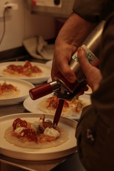 A person is using a culinary siphon to add a cream-like substance to a dish. The plate consists of thinly sliced rounds, likely carpaccio, topped with slices of meats, herbs, and dollops of cream. The setting appears to be a kitchen with additional similar plates in the background, indicating a food preparation or plating process.