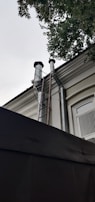 Two large metallic pipes extend vertically from a building, leading up to the roof where they are capped with black covers. A metal ladder is attached to the side of the building providing access to the roof and pipes. The building has a window with white bars and is partially obscured by a tree with green leaves at the top right corner of the image.