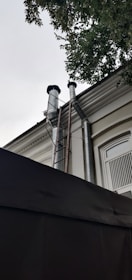 Two large metallic pipes extend vertically from a building, leading up to the roof where they are capped with black covers. A metal ladder is attached to the side of the building providing access to the roof and pipes. The building has a window with white bars and is partially obscured by a tree with green leaves at the top right corner of the image.