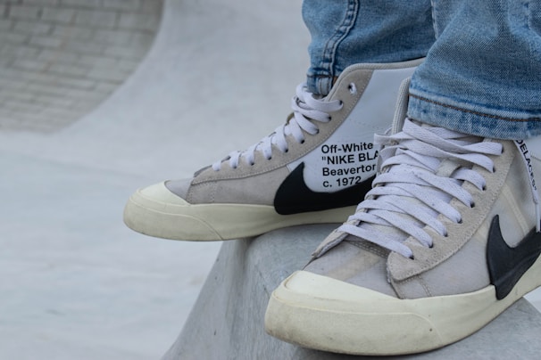 A close-up of a person's feet wearing high-top sneakers. The sneakers are a collaboration between Off-White and Nike, featuring a beige and white color scheme with black detailing and text on the side. The shoes rest on a smooth, curved concrete surface, possibly part of a skate park. The person is dressed in light blue jeans.