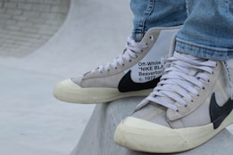 A close-up of a person's feet wearing high-top sneakers. The sneakers are a collaboration between Off-White and Nike, featuring a beige and white color scheme with black detailing and text on the side. The shoes rest on a smooth, curved concrete surface, possibly part of a skate park. The person is dressed in light blue jeans.