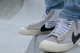A close-up of a person's feet wearing high-top sneakers. The sneakers are a collaboration between Off-White and Nike, featuring a beige and white color scheme with black detailing and text on the side. The shoes rest on a smooth, curved concrete surface, possibly part of a skate park. The person is dressed in light blue jeans.