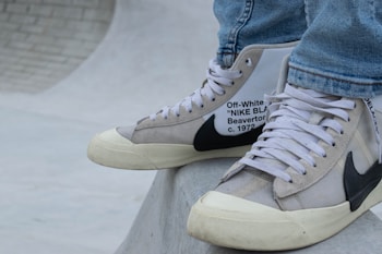 A close-up of a person's feet wearing high-top sneakers. The sneakers are a collaboration between Off-White and Nike, featuring a beige and white color scheme with black detailing and text on the side. The shoes rest on a smooth, curved concrete surface, possibly part of a skate park. The person is dressed in light blue jeans.