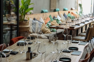 A large, wooden dining table is set with empty wine glasses and neatly placed napkins. The table extends alongside a cozy booth lined with colorful cushions, featuring shades of green, orange, and beige. Potted plants and wicker chairs add a touch of natural decoration, enhancing the inviting atmosphere.