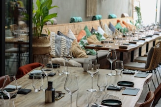 A large, wooden dining table is set with empty wine glasses and neatly placed napkins. The table extends alongside a cozy booth lined with colorful cushions, featuring shades of green, orange, and beige. Potted plants and wicker chairs add a touch of natural decoration, enhancing the inviting atmosphere.