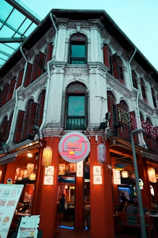 A traditional building with detailed architectural elements houses a bustling restaurant. The facade features intricate columns and windows with wooden shutters. The entrance is adorned with colorful, illuminated signs and lanterns, creating a vibrant and inviting atmosphere. Several people are seen dining inside, and menu boards are displayed at the front.
