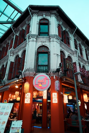 A traditional building with detailed architectural elements houses a bustling restaurant. The facade features intricate columns and windows with wooden shutters. The entrance is adorned with colorful, illuminated signs and lanterns, creating a vibrant and inviting atmosphere. Several people are seen dining inside, and menu boards are displayed at the front.