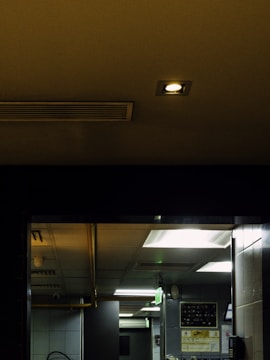 A dimly lit interior space featuring a ceiling with recessed lighting and ventilation. The room includes several overhead fluorescent lights emitting a greenish glow, and walls with various equipment. Posters or instructional sheets are visible on the wall, and a fire escape sign hangs from the ceiling.