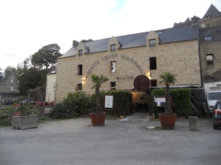 A rustic stone building with a sign reading 'Theatre Sarah Bernhardt' and 'Roche'. The building has a slate roof and multiple small windows. In front of it, there are two large potted palm trees, some greenery, and a seating area with red benches. A gray car is parked to the right side.
