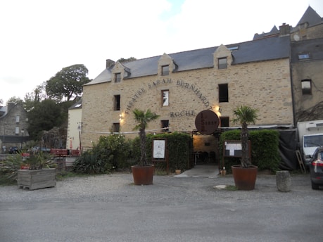 A rustic stone building with a sign reading 'Theatre Sarah Bernhardt' and 'Roche'. The building has a slate roof and multiple small windows. In front of it, there are two large potted palm trees, some greenery, and a seating area with red benches. A gray car is parked to the right side.
