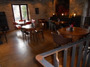 A cozy indoor café with wooden tables and a variety of chairs. The interior features stone walls and a wooden floor, creating a rustic vibe. There are several small tables, each set with glass jars containing utensils and napkins. Natural light comes in through a window on the left. The ambience is warm with dim lighting from candles on some tables.