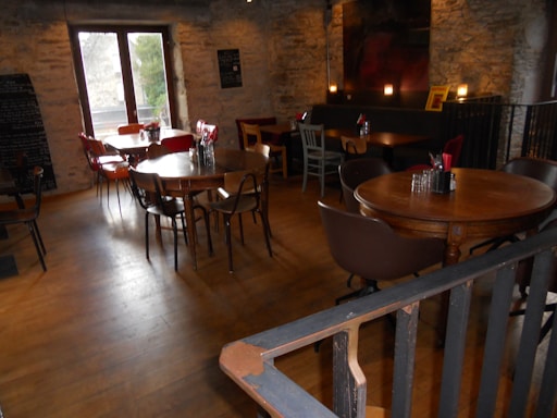 A cozy indoor café with wooden tables and a variety of chairs. The interior features stone walls and a wooden floor, creating a rustic vibe. There are several small tables, each set with glass jars containing utensils and napkins. Natural light comes in through a window on the left. The ambience is warm with dim lighting from candles on some tables.