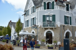 A charming, rustic building with stone walls and green shutters, identified as a hotel. It features a small balcony and an outdoor seating area with umbrellas. People walk by casually, giving a sense of leisure and relaxation. The sky is partly cloudy, adding to the pleasant atmosphere.