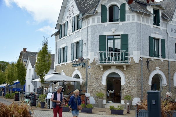 A charming, rustic building with stone walls and green shutters, identified as a hotel. It features a small balcony and an outdoor seating area with umbrellas. People walk by casually, giving a sense of leisure and relaxation. The sky is partly cloudy, adding to the pleasant atmosphere.