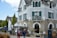 A charming, rustic building with stone walls and green shutters, identified as a hotel. It features a small balcony and an outdoor seating area with umbrellas. People walk by casually, giving a sense of leisure and relaxation. The sky is partly cloudy, adding to the pleasant atmosphere.