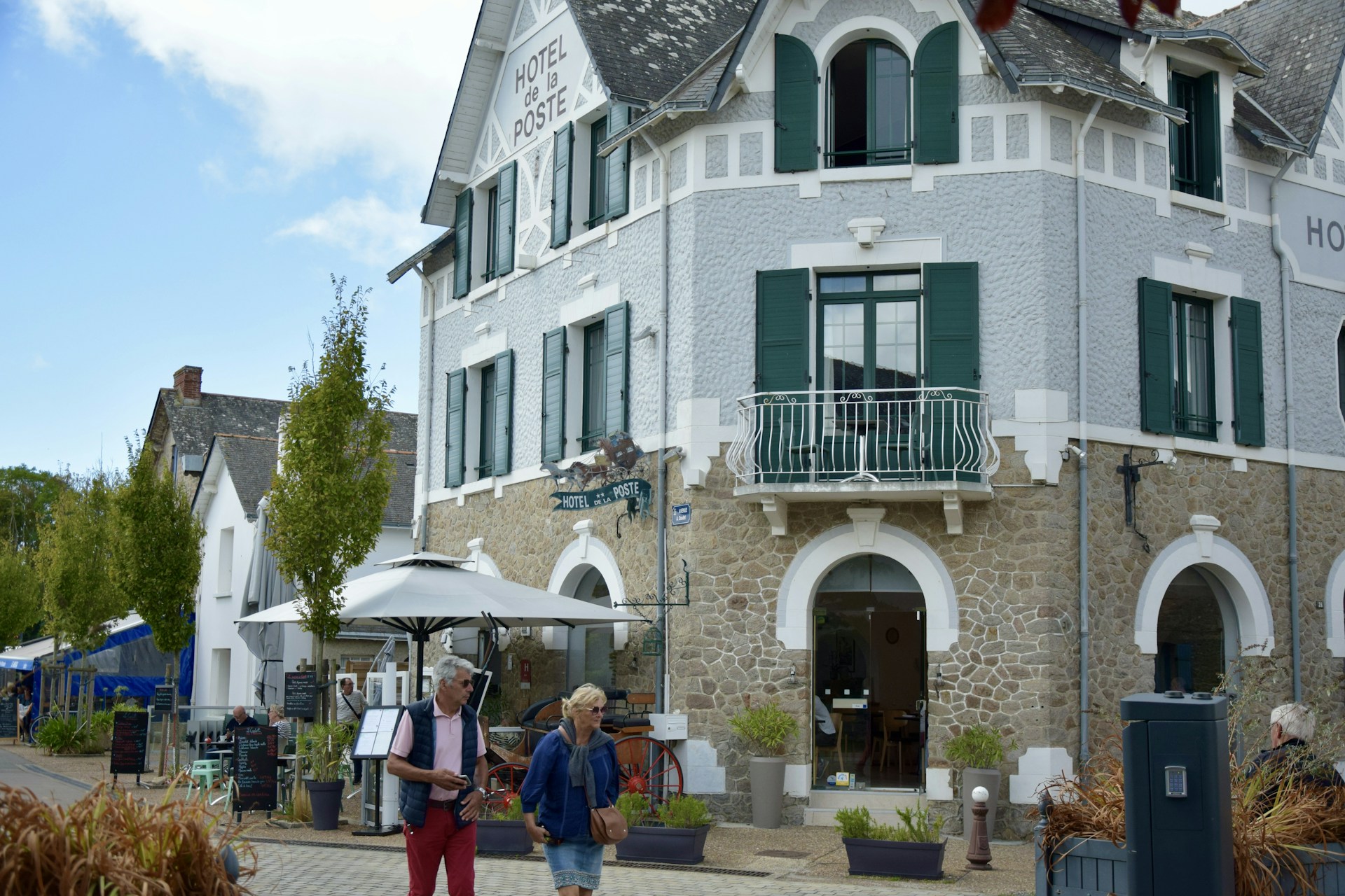 A charming, rustic building with stone walls and green shutters, identified as a hotel. It features a small balcony and an outdoor seating area with umbrellas. People walk by casually, giving a sense of leisure and relaxation. The sky is partly cloudy, adding to the pleasant atmosphere.