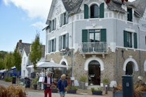 A charming, rustic building with stone walls and green shutters, identified as a hotel. It features a small balcony and an outdoor seating area with umbrellas. People walk by casually, giving a sense of leisure and relaxation. The sky is partly cloudy, adding to the pleasant atmosphere.