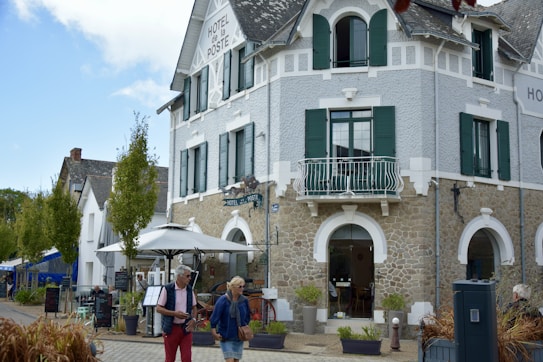 A charming, rustic building with stone walls and green shutters, identified as a hotel. It features a small balcony and an outdoor seating area with umbrellas. People walk by casually, giving a sense of leisure and relaxation. The sky is partly cloudy, adding to the pleasant atmosphere.
