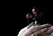 An elderly man wearing a dark jacket and cap is deeply focused on sewing or stitching a large piece of fabric. He is using a needle and thread, creating an impression of careful craftsmanship. The background is dark, emphasizing the man and his work in the spotlight.
