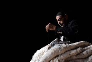 An elderly man wearing a dark jacket and cap is deeply focused on sewing or stitching a large piece of fabric. He is using a needle and thread, creating an impression of careful craftsmanship. The background is dark, emphasizing the man and his work in the spotlight.