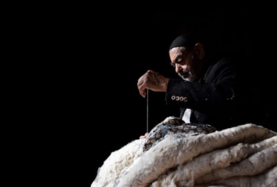 An elderly man wearing a dark jacket and cap is deeply focused on sewing or stitching a large piece of fabric. He is using a needle and thread, creating an impression of careful craftsmanship. The background is dark, emphasizing the man and his work in the spotlight.