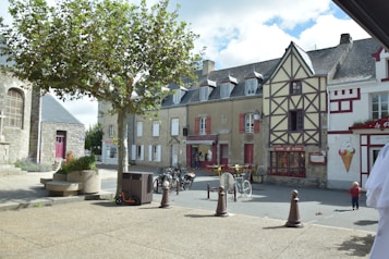 A quaint European street scene features a mix of traditional stone and half-timbered buildings, with a large tree providing shade over a paved area. Bicycles are parked near a caf&eacute; with outdoor seating, showing a few people enjoying the ambience. The scene also includes an ice cream shop with a large cone painted on the wall. A toddler is visible in the foreground exploring the space.